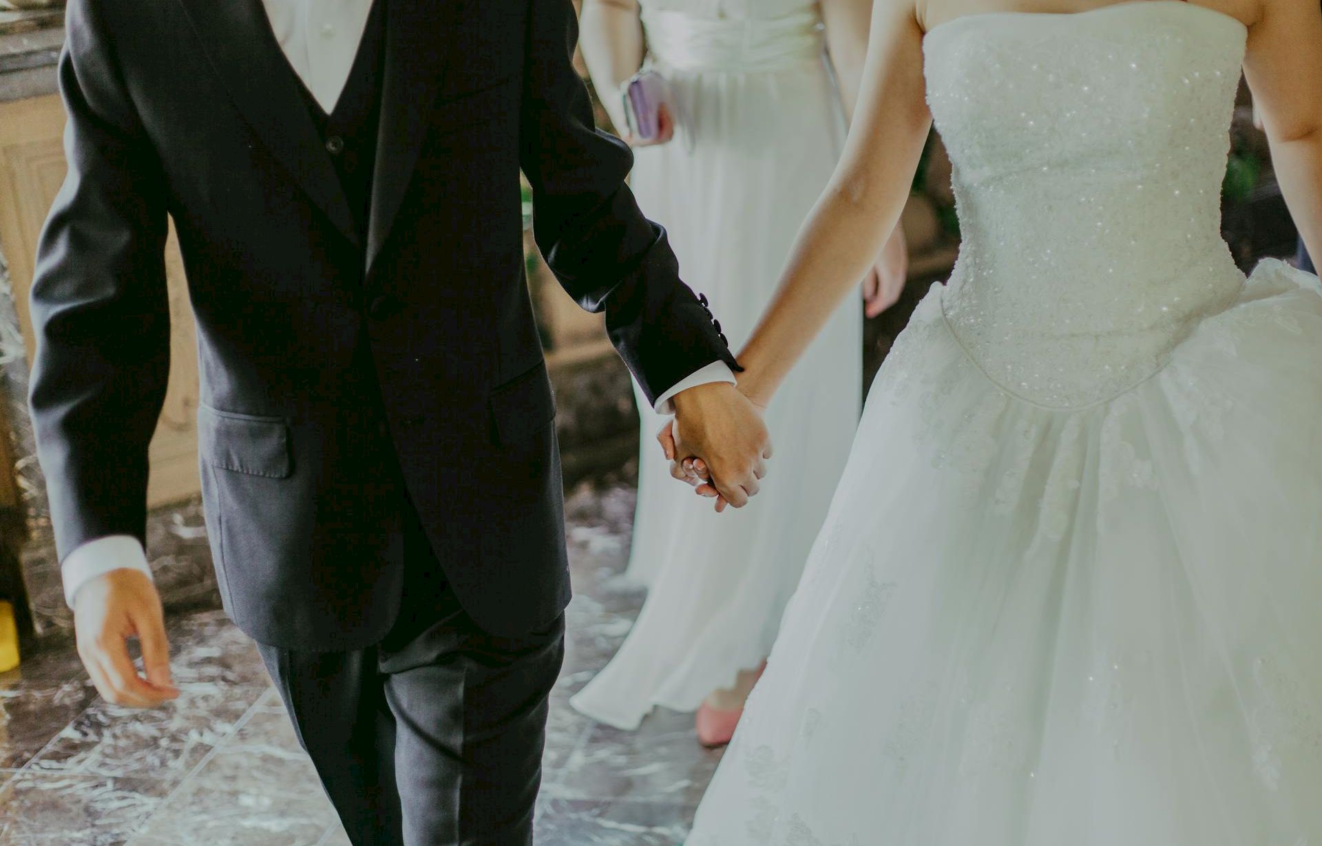 A couple dressed in wedding attire&mdash;groom in a black suit and tie, bride in a white wedding gown, holding hands as they walk together.