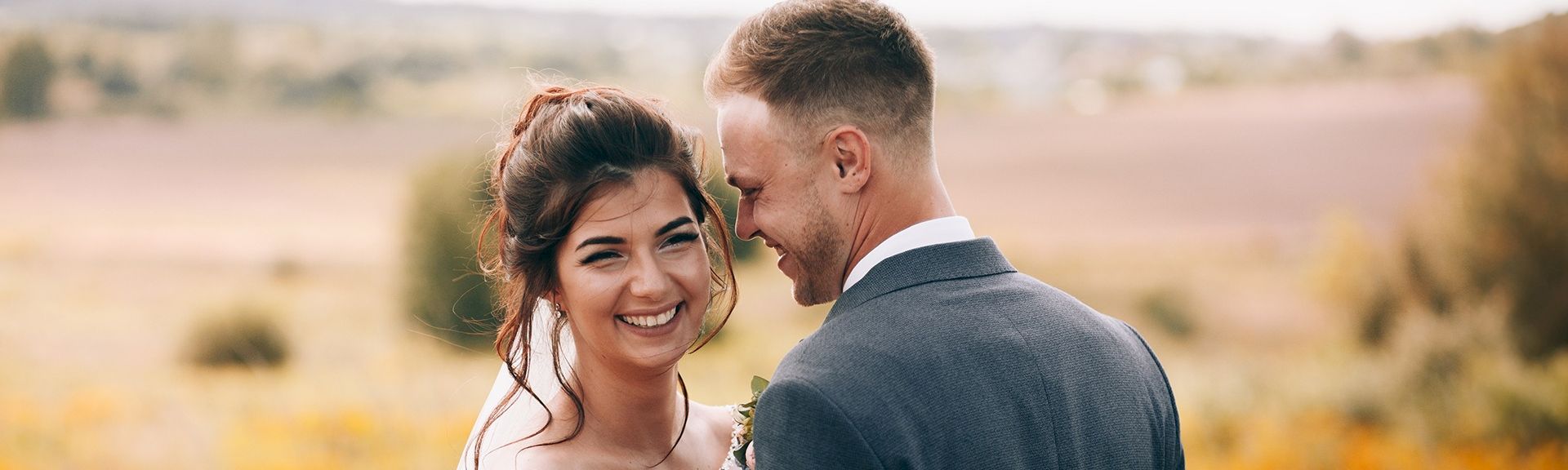 Two smiling people in a joyful embrace outdoors, a couple dressed nicely with a warm, sunlit field behind them.
