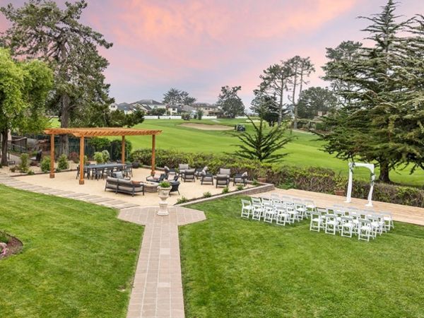Outdoor wedding setup on a lawn with white chairs, a wooden pergola, paved paths, and a scenic golf-like green in the background, at sunset.