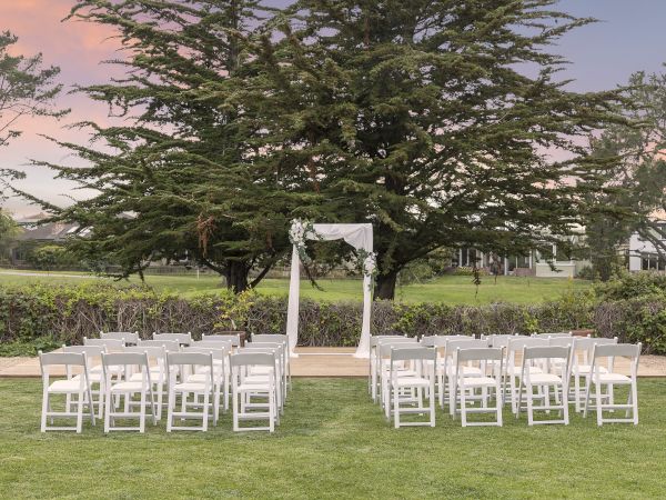 An outdoor wedding setup with a white arch, rows of white chairs on a grassy lawn, and a large tree in the background, ready for ceremony.