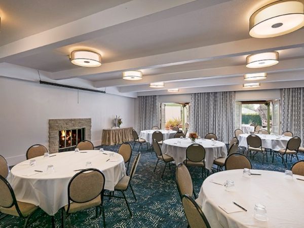 A formal banquet room with round tables, white tablecloths, cushioned chairs, soft lighting, and a long sideboard near windows with patterned curtains.