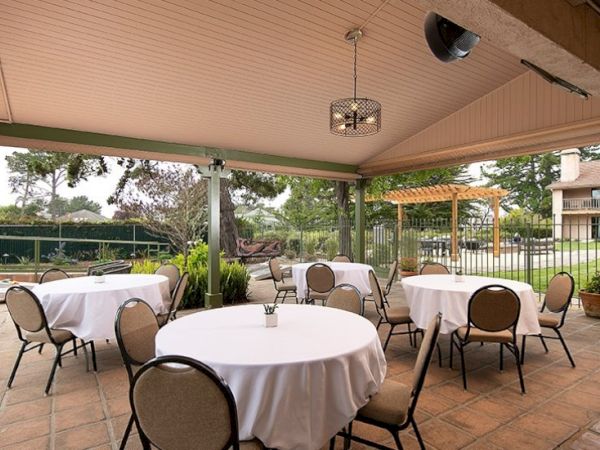 An empty outdoor restaurant patio with round tables draped in white cloths, beige chairs, and potted plants under a covered ceiling.