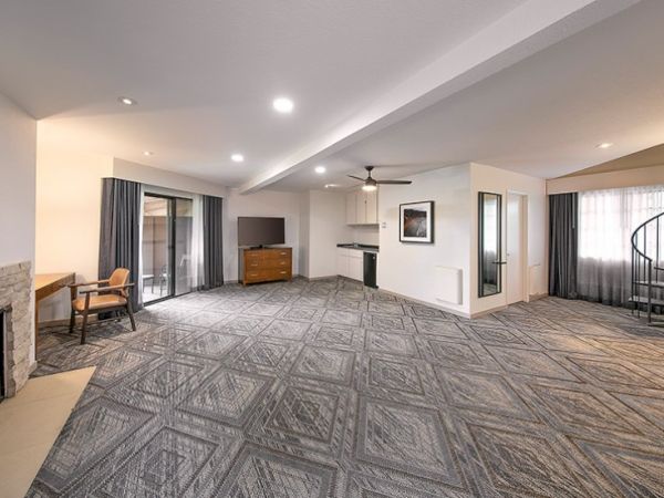 A spacious, modern living area with patterned carpet, a fireplace, a small dining nook, and a balcony door, plus a corner desk and TV setup, all in soft lighting.