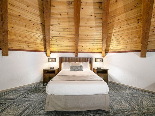 A cozy attic bedroom with a central bed, wooden beams overhead, two nightstands, lamps, neutral linens, and a patterned carpet.