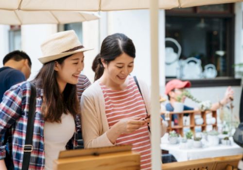 Two friends are shopping at an outdoor market, smiling and looking at a phone together while browsing items under an umbrella.