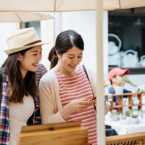 Two friends are shopping at an outdoor market, smiling and looking at a phone together while browsing items under an umbrella.