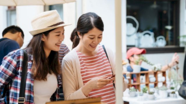 Two friends are shopping at an outdoor market, smiling and looking at a phone together while browsing items under an umbrella.