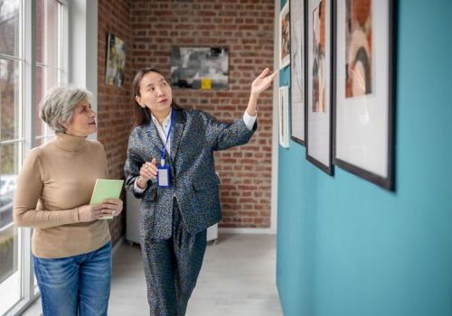 Two women look at framed photos in a bright hallway; one explains while pointing, the other holds a notebook and listens attentively.