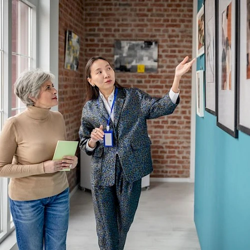 Two women look at framed photos in a bright hallway; one explains while pointing, the other holds a notebook and listens attentively.