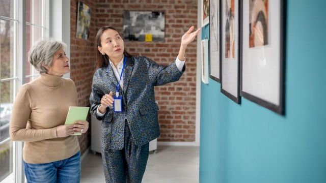 Two women look at framed photos in a bright hallway; one explains while pointing, the other holds a notebook and listens attentively.