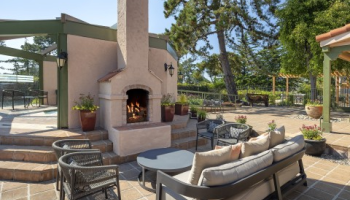 A sunny outdoor patio with a stone fireplace, beige cushioned seating, black wicker chairs, potted plants, and a tidy tiled deck under trees, inviting relaxation.