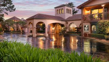 A Mediterranean-style hotel with arches, tower, and warm lights; a reflective pool in front, lush greenery, and pink sunset skies.