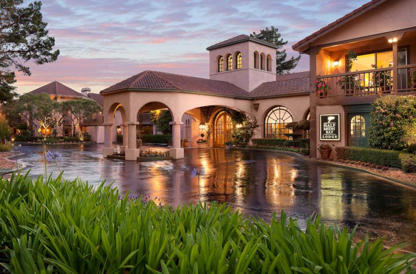 A Mediterranean-style hotel with arches, tower, and warm lights; a reflective pool in front, lush greenery, and pink sunset skies.