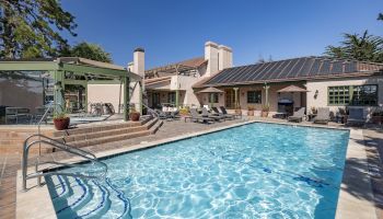A sunny backyard pool scene with crystal blue water, a surrounding tiled deck, lounge chairs, a pergola, and a beige house with solar panels in the background.