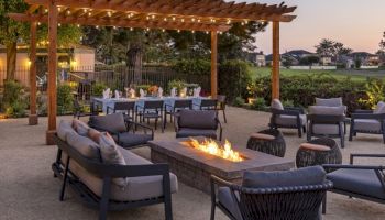 Outdoor lounge with a wooden pergola, comfy couches, fire pit, string lights, and a dining table setup on a paved patio near a green field.