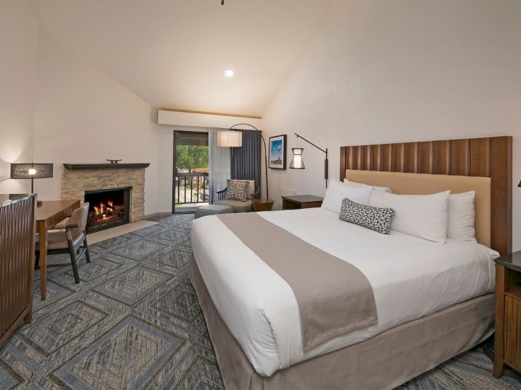 A hotel room with a large bed, nightstands, a fireplace, a desk, and a seating area; sliding door to a balcony and a calm, modern decor.