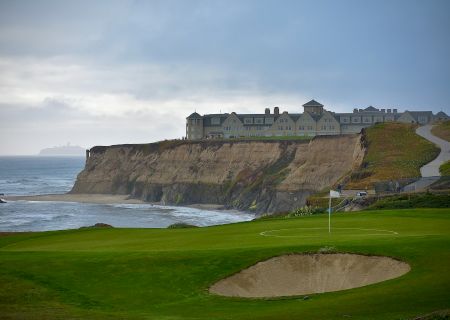 Cliffside with a coastal golf course, sand bunker, green fairways, and a coastal fortress-style building perched above the sea under a gray sky.