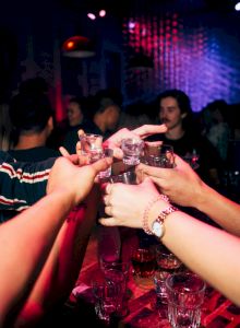 A group of people clinking shot glasses over a table in a dim, colorful club setting, cheering together to celebrate.