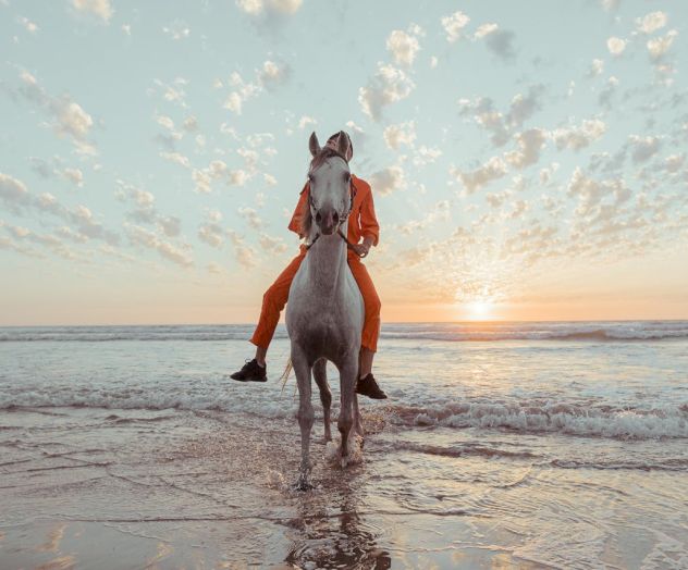 A person rides a white horse into shallow surf at sunset, with orange pants and a peaceful, cloudy sky above the calm sea.