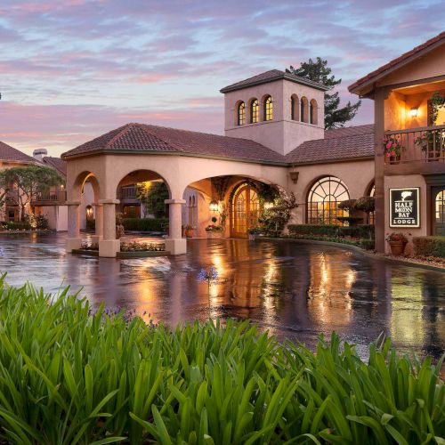 A luxurious resort with a Spanish-style building, arches, and a reflective pool at sunset, surrounded by greenery and warm lighting.