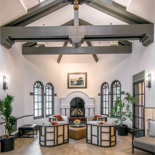 A bright, elegant living room with high ceilings, exposed beams, arched windows, a fireplace, and modern seating around a central table.