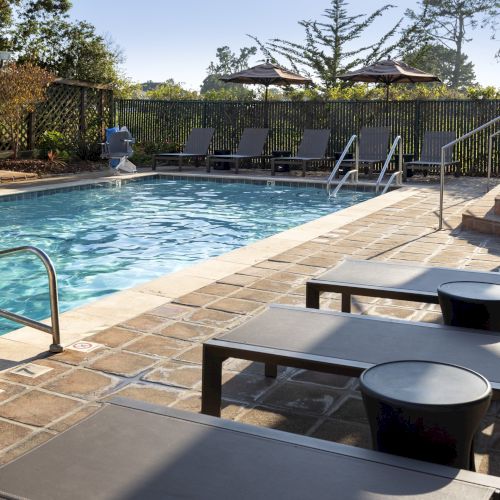 An outdoor pool area with sun loungers, small side tables, and a fence; clear water, paved deck, and trees in the background.