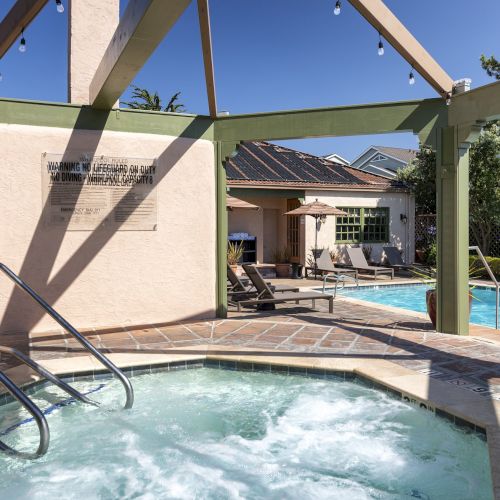 A relaxed pool area with a hot tub in the foreground, a sunny resort pool, lounge chairs, and a shaded seating area under a wooden pergola.