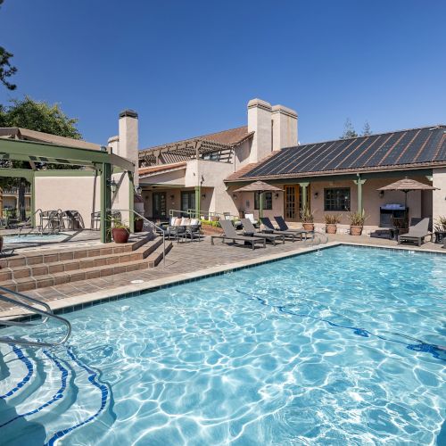 A sunny backyard with a rectangular in-ground pool, blue water, tiled deck, lounge chairs, and a beige house with solar panels surrounding the area.