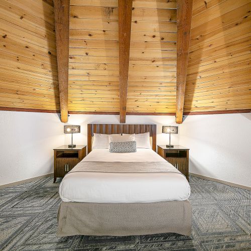 A cozy attic bedroom with a centered bed, wooden beam ceiling, two nightstands, lamps, and a patterned rug, neatly arranged in warm tones.