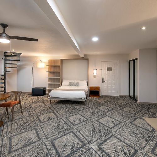 A modern hotel suite with a bed, sofa, table and chairs, a fireplace, and a spiral staircase leading to a loft area; bright lighting throughout.