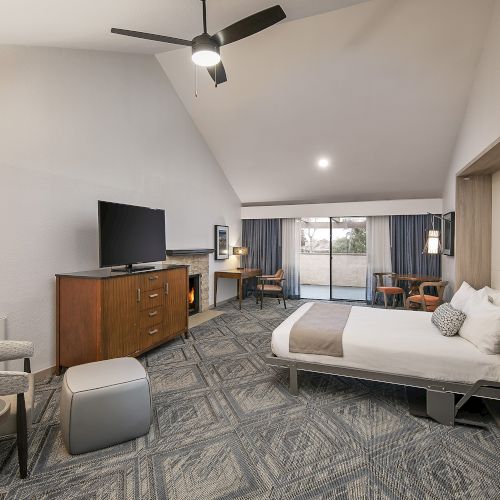 A spacious hotel room with a king bed, desk, TV on a dresser, a seating area with chairs and a round table, a ceiling fan, and open shelving along one wall.