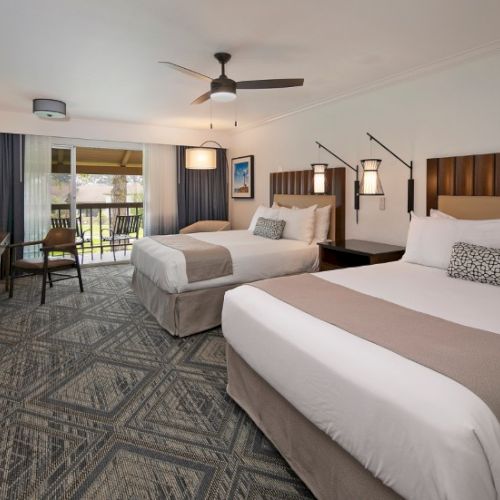 Two beds in a modern hotel room with a balcony, soft beige and gray tones, a desk, chairs, and ceiling fan.