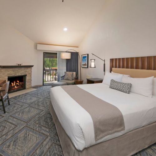 A hotel room with a large bed, fireplace, desk, sitting area, and a balcony access, all in a cozy, neutral-toned suite.