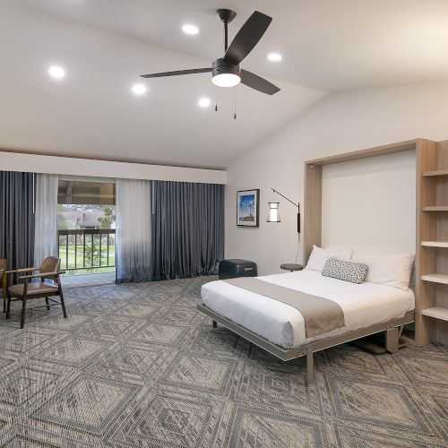 A hotel room with a king bed, desk, TV, open shelves, and a seating area near sliding glass doors with blue curtains, ceiling fan, and patterned carpet.