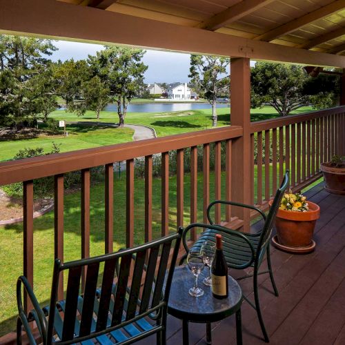Cozy balcony overlooking a sunny lawn with trees, a winding path, and a water feature in the distance; a table with drinks and chairs awaits.