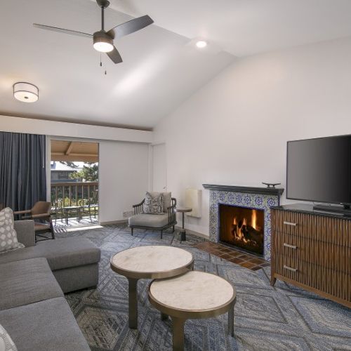 A cozy living room with a gray sectional, two round coffee tables, a fireplace, a large flat-screen TV on a wooden cabinet, and a ceiling fan with lights.