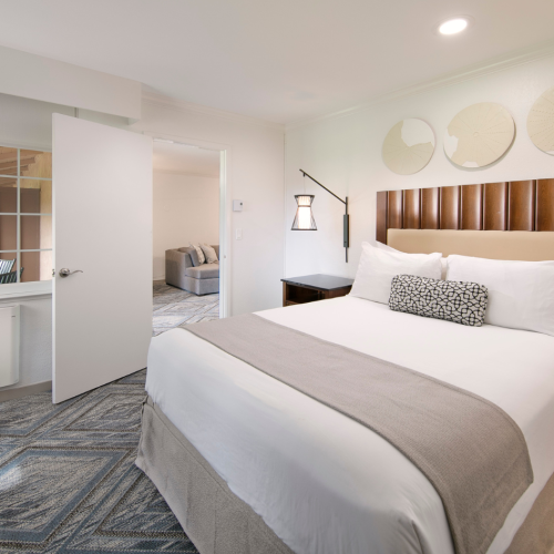 A modern hotel room with a king bed, wooden headboard, neutral linens, two nightstands, pendant lamps, and a patterned rug; a doorway leads to a seating area.
