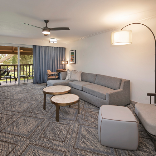 Spacious living room with a gray sectional, two round coffee tables, a matching ottoman, a curved floor lamp, and a patterned blue-gray rug; sliding doors lead to a balcony.
