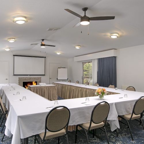 A conference room with a U-shaped table setup, white tablecloths, beige chairs, a projector screen, a fireplace, ceiling fans, and blue carpet in a bright, tidy space.