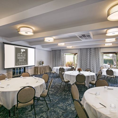 A conference room set up for a banquet or meeting with round tables, white tablecloths, place settings, and a screen displaying &ldquo;Hall & Lodge.&rdquo;