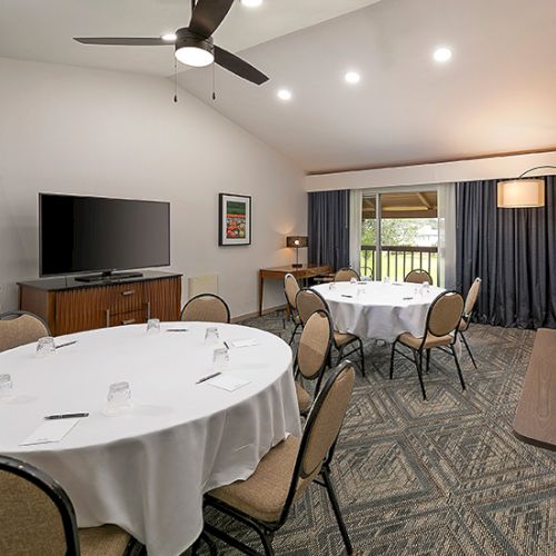 A conference room or meeting space with round tables, chairs, a large TV, whiteboard, and a coffee bar area, set up for a presentation or event.