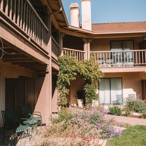 A two-story brown apartment building with balconies, a manicured garden bed, climbing vines, and outdoor chairs by the walkway.