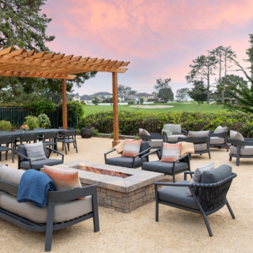 A stylish outdoor lounge area with cushioned seating around a central fire pit, under a wooden pergola, with trees and a scenic backdrop.