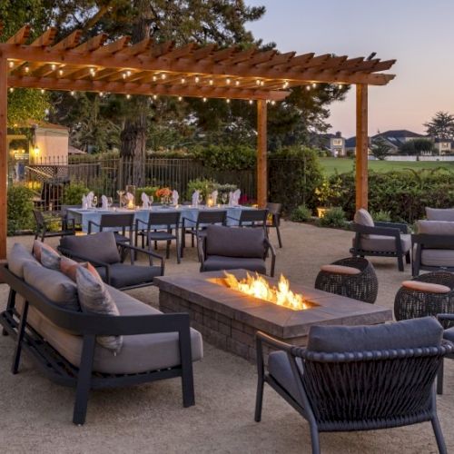 Outdoor lounge area with a wooden pergola, sectional sofas, lounge chairs, and a long dining table; a fire pit glows warmly at dusk.