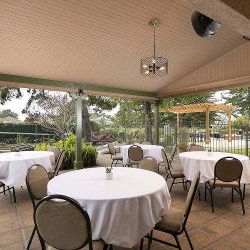 A covered outdoor dining area with round tables, white tablecloths, and beige chairs set for guests, on a patio with greenery nearby.