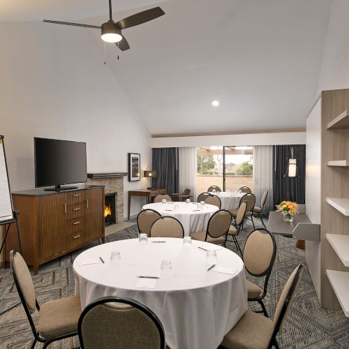 A bright conference room with round tables, beige chairs, a wall-mounted TV, a whiteboard, and a ceiling fan in a quiet meeting space.