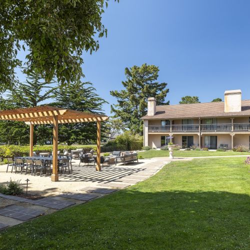 A sunny outdoor courtyard with a lawn, a wooden pergola-covered seating area, paved paths, and a two-story building in the background, perfect for relaxing.