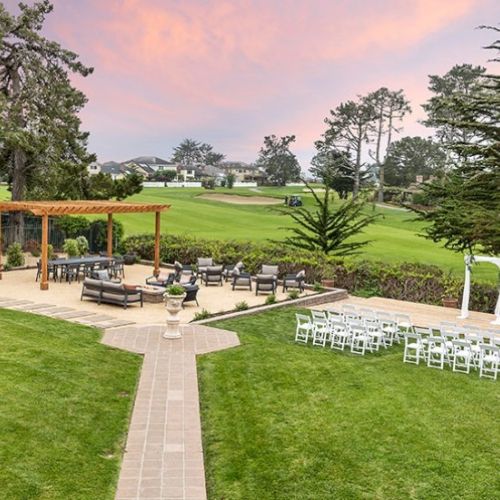 An outdoor wedding setup on a well-kept lawn with a wooden pergola, chairs arranged in rows, a paved path, and scenic trees under a pink-tinged sky.