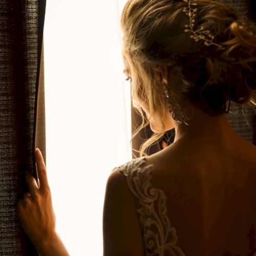 A woman in a dress with an intricate back design stands by a window, her hair styled up and hands on the curtain, facing the light.