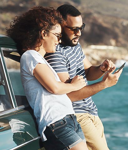 A couple leaning against a teal vintage car by the coast, looking at a tablet together as waves roll in the background.